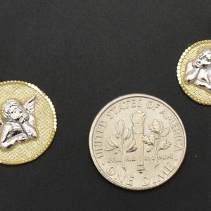 May include: Two gold angel pendant charms with a white angel design. The charms are shown next to a US dime for size comparison.