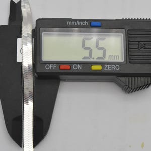Puede incluir: Una cadena de plata en forma de espiga se mide con un calibrador digital. La pantalla digital muestra 5,5 mm. La cadena es plana y tiene una superficie reflectante. El calibrador es negro.