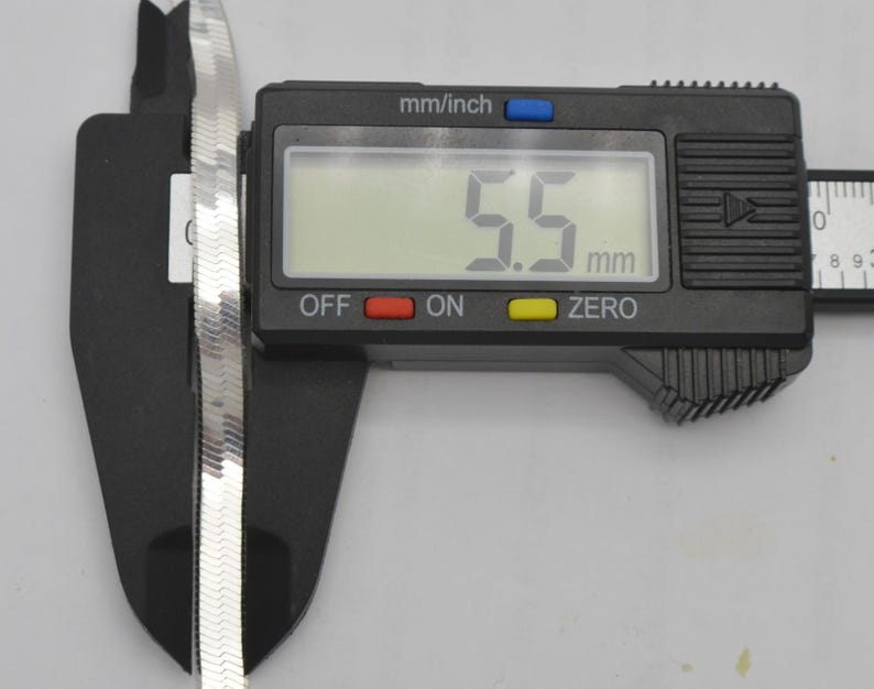 Puede incluir: Una cadena de plata en forma de espiga se mide con un calibrador digital. La pantalla digital muestra 5,5 mm. La cadena es plana y tiene una superficie reflectante. El calibrador es negro.