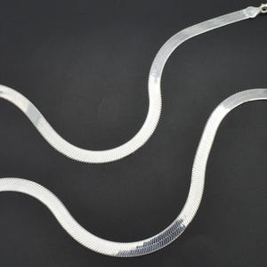 Puede incluir: Un collar de cadena plateada con dise&ntilde;o de espiga. El collar es plano y flexible, con una superficie lisa y reflectante. La cadena se muestra sobre un fondo negro, resaltando su brillo met&aacute;lico. Una pieza de joyer&iacute;a cl&aacute;sica.