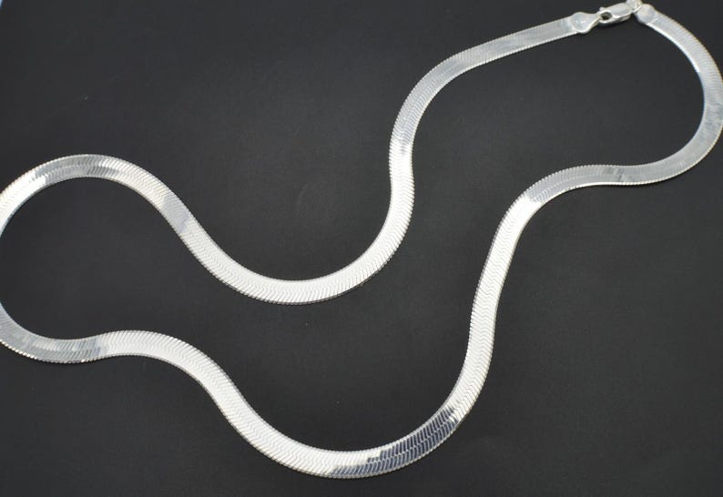 Puede incluir: Un collar de cadena plateada con dise&ntilde;o de espiga. El collar es plano y flexible, con una superficie lisa y reflectante. La cadena se muestra sobre un fondo negro, resaltando su brillo met&aacute;lico. Una pieza de joyer&iacute;a cl&aacute;sica.