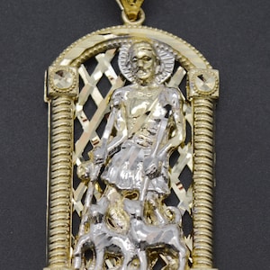 May include: A gold pendant featuring a detailed image of Saint Lazarus, a religious figure, with two dogs. The pendant is made of two-tone gold and has a diamond-shaped lattice design. The inscription "SAINT LAZARUS" is visible at the bottom.