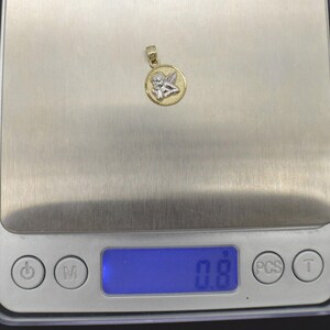 May include: A gold and silver pendant featuring a cherub angel design. The pendant is on a digital scale that reads 0.8.