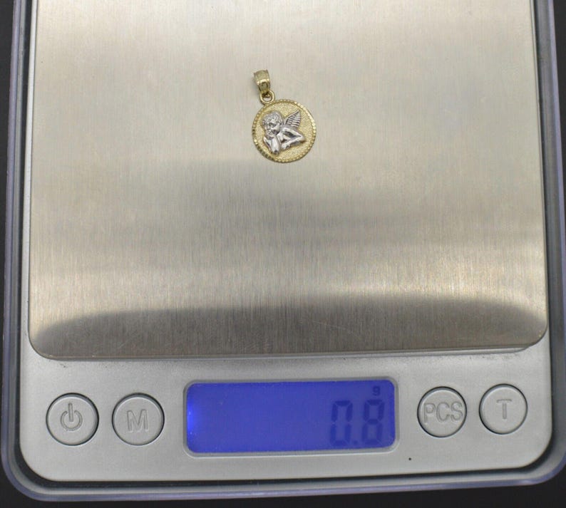 May include: A gold and silver pendant featuring a cherub angel design. The pendant is on a digital scale that reads 0.8.