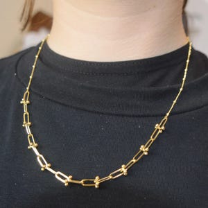 May include: A gold-toned necklace featuring a series of elongated rectangular links connected by small spherical beads. The chain transitions into a delicate, thin cable chain at the upper sections, resting against a plain black fabric background.