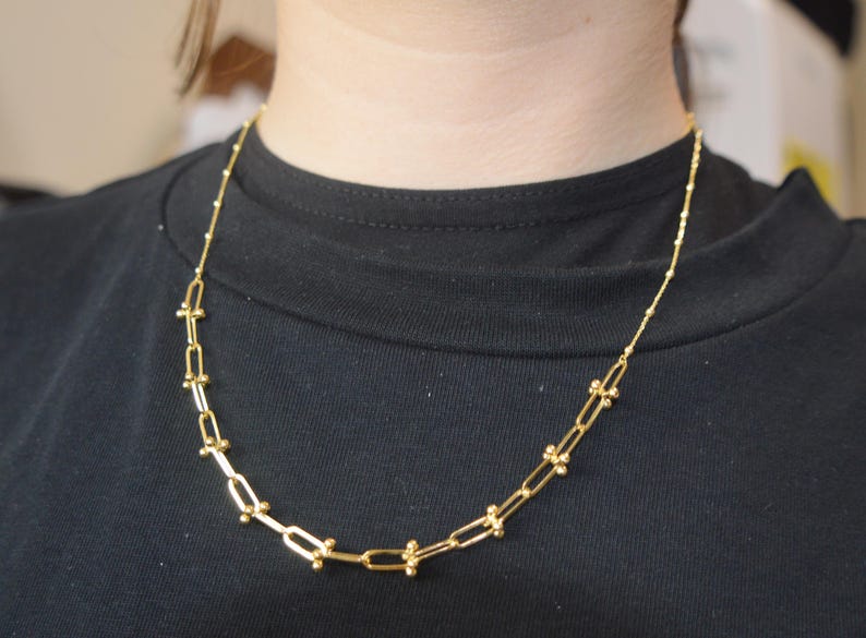 May include: A gold-toned necklace featuring a series of elongated rectangular links connected by small spherical beads. The chain transitions into a delicate, thin cable chain at the upper sections, resting against a plain black fabric background.