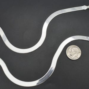 Puede incluir: Un collar de cadena plateada en espiga con cierre, sobre una superficie negra. Una moneda est&aacute; al lado del collar para comparar el tama&ntilde;o. La cadena tiene un dise&ntilde;o plano y tejido.
