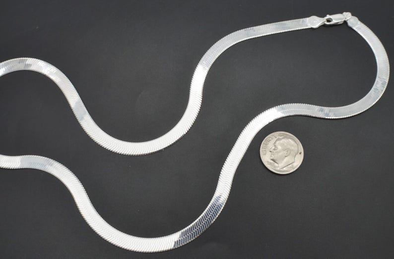 Puede incluir: Un collar de cadena plateada en espiga con cierre, sobre una superficie negra. Una moneda est&aacute; al lado del collar para comparar el tama&ntilde;o. La cadena tiene un dise&ntilde;o plano y tejido.