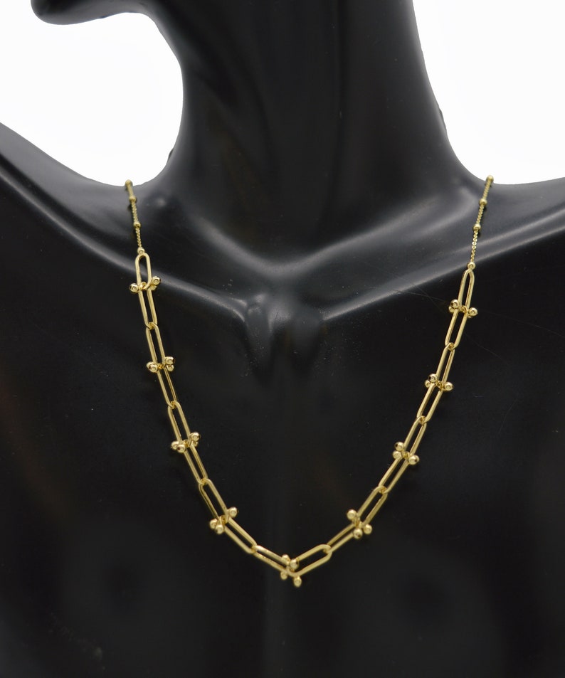 May include: A gold-toned chain necklace featuring elongated oval links connected by small, spherical beads at each joint. The necklace has a delicate, uniform structure with a polished finish, creating a continuous, repeating pattern of links and beads.