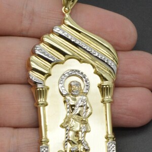 May include: Gold pendant featuring a religious figure with a halo, staff, and sheep. The pendant has a gold and silver-tone design with an arched top and decorative details. A stylish piece of jewelry.