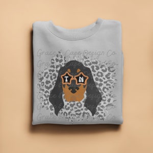 Puede incluir: Una sudadera gris con un dachshund negro y marrón que lleva gafas de sol con las letras "TN" en ellas. El dachshund está rodeado de un estampado de leopardo blanco y gris.