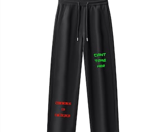 May include: Black sweatpants with a drawstring waist. The text "WILD & FREE" is printed in red on the left leg, and "CAN'T TAME HER" is printed in green on the right leg. The pants have a relaxed fit.