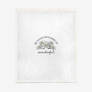 May include: White throw blanket featuring a daisy floral design and the text "Be your own kind of wonderful" in black script. The blanket has a soft texture and a white border.