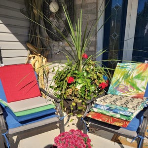 May include: Two dark metal chairs holding stacks of square seat cushions. The left stack features solid red, grey, green, and blue cushions. The right stack displays cushions with tropical leaf, geometric, floral, and red botanical patterns.
