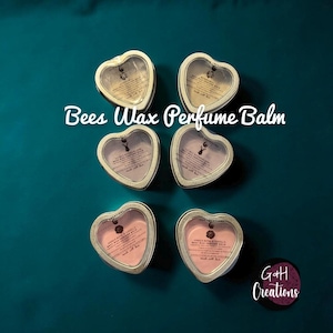 May include: Six heart-shaped Bees Wax Perfume Balm tins with a clear top, arranged on a teal background. The tins have a silver rim and contain different colored balms. The text "Bees Wax Perfume Balm" is written across the image.