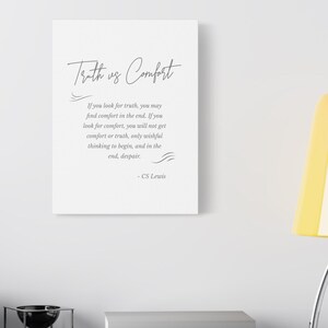 Peut inclure: Une impression sur toile blanche avec l'inscription "Truth vs Comfort" en écriture cursive. En dessous, une citation de C.S. Lewis est imprimée dans une police claire. L'impression est affichée sur une surface grise avec des livres et une lampe.