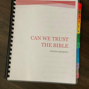 May include: A white binder with a red, white, and blue striped cover. The title "CAN WE TRUST THE BIBLE" is printed in black on the cover. The subtitle "Christian Apologetics" is printed in black below the title. The binder has a black plastic spiral binding and colorful page dividers.