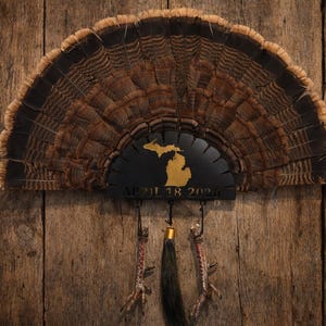 May include: A turkey fan display with a black metal accent featuring a gold-colored outline of the state of Michigan and the date "APRIL 18 2026". The fan is mounted on a wooden background.