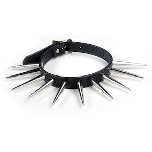 May include: A black leather choker collar with silver metal spikes. The collar features a buckle fastening. The spikes are evenly spaced around the collar, designed to be worn around the neck.