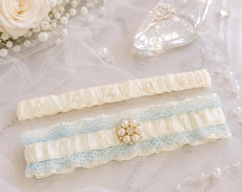 May include: A set of two ivory satin garters. The wider garter features light blue lace trim and a central floral-shaped embellishment made of faux pearls and clear crystals. The narrower garter is accented with small, evenly spaced faux pearls.