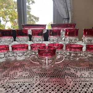 May include: A collection of clear glass drinkware with a ruby red rim. The set includes teacups with handles, small glasses, and serving bowls. The glass has a textured pattern. The set is arranged on a white lace tablecloth.