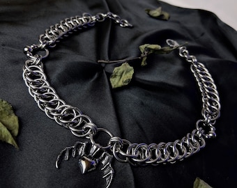 May include: A silver chain necklace with a pendant featuring a heart and bat wings. The necklace is on a black fabric background.