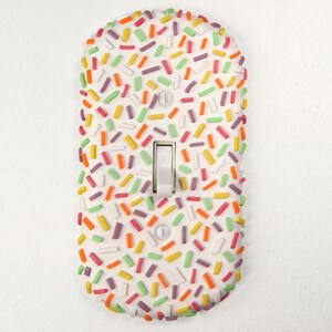 Könnte beinhalten: Ein weißer Lichtschalter mit einem bunten Streusel-Design. Die Streusel sind in verschiedenen Schattierungen von Rosa, Grün, Orange und Gelb.