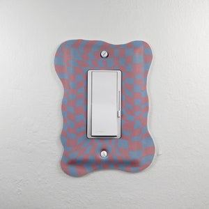 Könnte beinhalten: Ein weißer Lichtschalter mit einer rosafarbenen und blauen karierten Abdeckung. Die Abdeckung hat eine wellige, unregelmäßige Form.