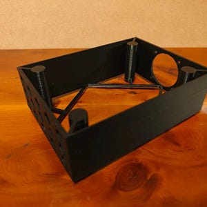 May include: Black plastic case with a circular opening and four cylindrical supports. The case is designed to hold a small electronic device.