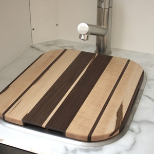 May include: A rectangular wooden cutting board with alternating stripes of light and dark wood. The board is sitting on a white marble countertop next to a stainless steel sink.