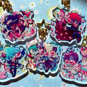 May include: Set of eight colorful character charms with glitter accents. Each charm features a different character with a unique design and is attached to a gold chain.