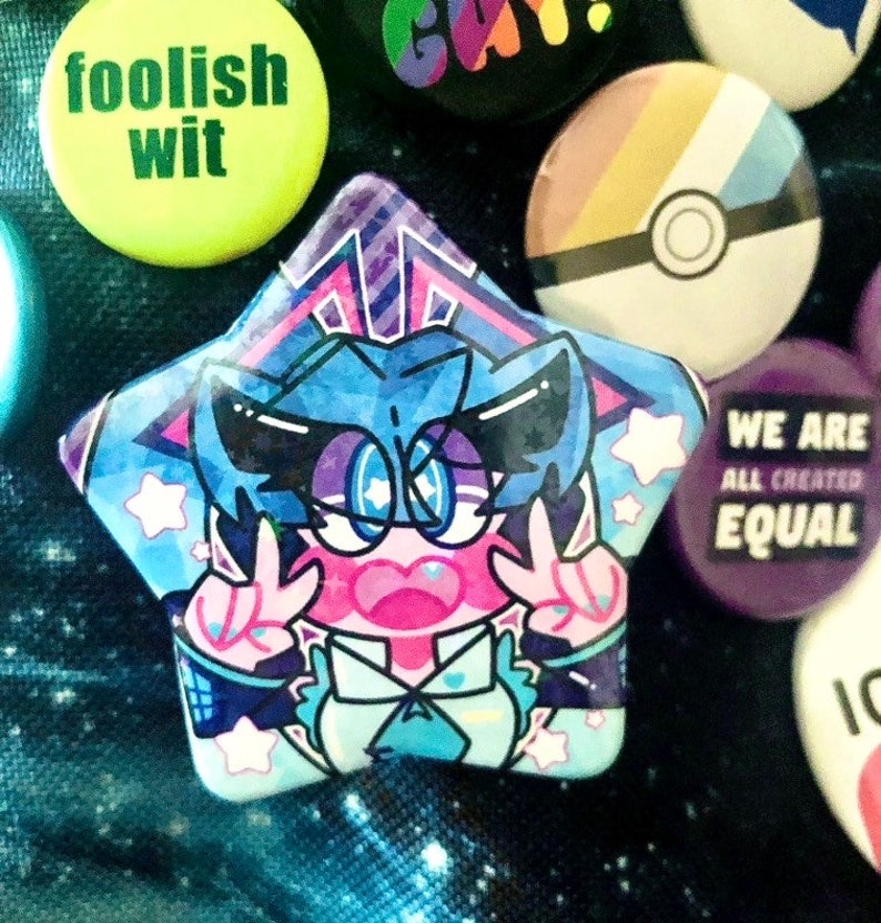 A star-shaped button pin of VOCALOID Hatsune Miku pinned to a blue galaxy-printed bag. She is accompanied by other buttons, mostly LGBTQ+ pride related, pinned to the bag on the right.