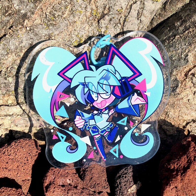 Peut inclure: Un personnage d'anime bleu et rose avec de longs cheveux bleus et un casque. Le personnage porte une tenue blanche et bleue et tient un microphone. Le personnage est entour&eacute; d'un fond bleu et rose avec des formes g&eacute;om&eacute;triques.