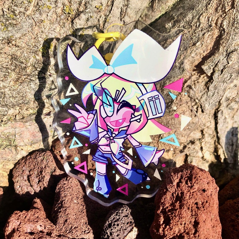 Peut inclure: Un porte-cl&eacute;s en acrylique transparent repr&eacute;sentant un personnage de style anime color&eacute; avec des cheveux roses et un n&oelig;ud bleu, portant une tenue bleu et blanche. L'arri&egrave;re-plan pr&eacute;sente un motif de triangles roses, bleus et blancs.