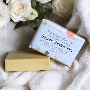 Può includere: Un sapone Secret Garden di The Witch & Cauldron, realizzato a mano nel Lancashire, con geranio rosa, neroli, lavanda e oli essenziali di ylang-ylang. Il sapone è di colore giallo chiaro e si trova su un asciugamano bianco con rose bianche.