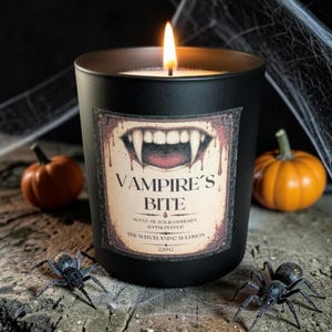 Puede incluir: Una vela negra mate en un frasco de vidrio con una mecha encendida. La etiqueta presenta un gráfico de colmillos de vampiro, el texto VAMPIRE'S BITE, SCENT: BLACK RASPBERRY & PINK PEPPER, THE WITCH AND CAULDRON y 220g. La vela está rodeada de pequeñas calabazas y arañas.