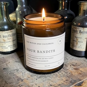 May include: A lit soy wax candle in an amber glass jar with a white label reading The Witch and Cauldron Four Bandits, Natural Essential Oil Soy Wax Candle, Hand-Poured in Lancashire/UK, Vegan/Cruelty Free, 170g, Burn 35h, surrounded by glass bottles.