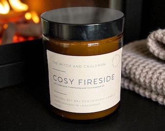 Op de afbeelding: Bruine glazen potkaars met een zwarte deksel. Het etiket luidt "COSY FIRESIDE". De kaars staat op een zwart oppervlak, met een open haard op de achtergrond. Handgegoten in Lancashire.