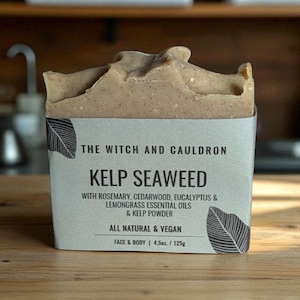 May include: A bar of Kelp Seaweed soap from The Witch and Cauldron, with a light brown, textured surface. The soap is wrapped in a gray label with the text "Kelp Seaweed" and ingredient details. The bar weighs 4.5 oz.