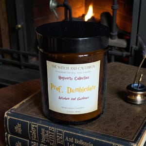 Peut inclure: Une bougie en pot de verre marron avec un couvercle noir. L'étiquette indique "THE WITCH AND CAULDRON" et "Prof. Dumbledore" de la "Hogwarts Collection". La bougie est sur une pile de vieux livres.