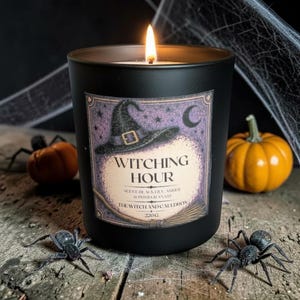 Op de afbeelding: Een matzwarte kaars in een glazen pot met een brandende lont. Het etiket heeft een paarse achtergrond, een zwarte heksenhoed, een maansikkel en de tekst "WITCHING HOUR, SCENT: BLACK LILY, AMBER & POMEGRANATE, THE WITCH AND CAULDRON, 220G".