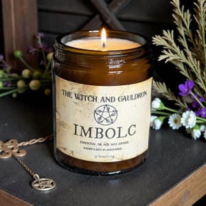 May include: A lit soy wax candle in a brown glass jar with a cream-colored label reading THE WITCH AND GAULDRON, IMBOLC, ESSENTIAL OIL SOY WAX CANDLE, HANDPOURED IN LANCASHIRE, 35 hour/150g, featuring a pentacle symbol in the center of the text.