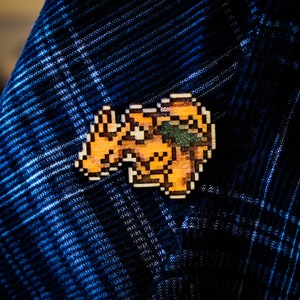 Op de afbeelding: Een gepixeld oranje en bruin ontwerp van een wezen, mogelijk een draak, is genaaid op een blauw-wit geruite stof.