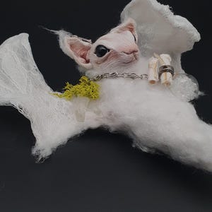 Pode incluir: Uma escultura artesanal de uma criatura fantástica com um rosto rosa pálido, orelhas grandes e olhos negros. Possui pelagem branca e fofa e delicadas asas rendadas. Adornada com uma corrente, cristais e um anel, sobre um fundo escuro.