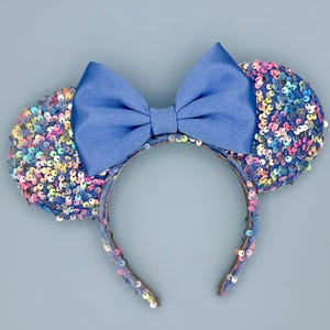 May include: Blue sequined Minnie Mouse ears with a blue satin bow. The ears are covered in iridescent sequins in shades of pink, blue, green, and purple.