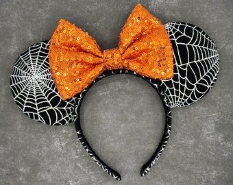 May include: Halloween-themed headband with black and white spiderweb ears and a large orange sequined bow. The headband is black with a white pattern. Perfect for Halloween costumes or themed events.