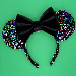 May include: Black headband with two round ears covered in colorful sequins. A large black velvet bow sits between the ears. The headband and ears are on a green background.