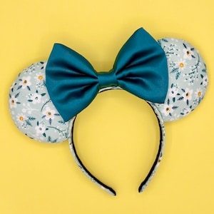May include: Teal bow headband with a floral print on the ears. The floral print is a light green with white flowers.