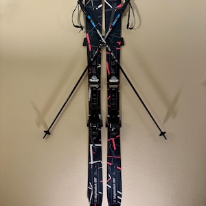 K&ouml;nnte beinhalten: Paar schwarzer und wei&szlig;er Ski mit rosa und wei&szlig;en geometrischen Designs, an einer Wand befestigt. Die Ski haben schwarze Bindungen und werden mit schwarzen Skist&ouml;cken kombiniert. Der Text "Nightstick 097" ist auf den Skiern sichtbar.