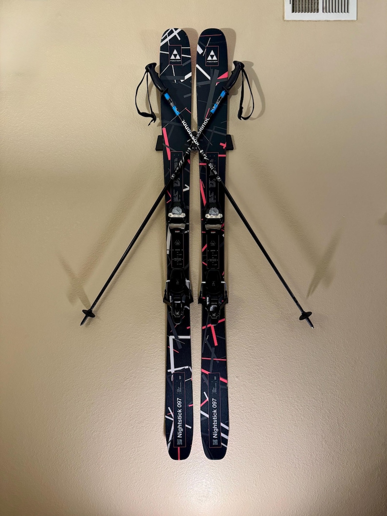 K&ouml;nnte beinhalten: Paar schwarzer und wei&szlig;er Ski mit rosa und wei&szlig;en geometrischen Designs, an einer Wand befestigt. Die Ski haben schwarze Bindungen und werden mit schwarzen Skist&ouml;cken kombiniert. Der Text "Nightstick 097" ist auf den Skiern sichtbar.
